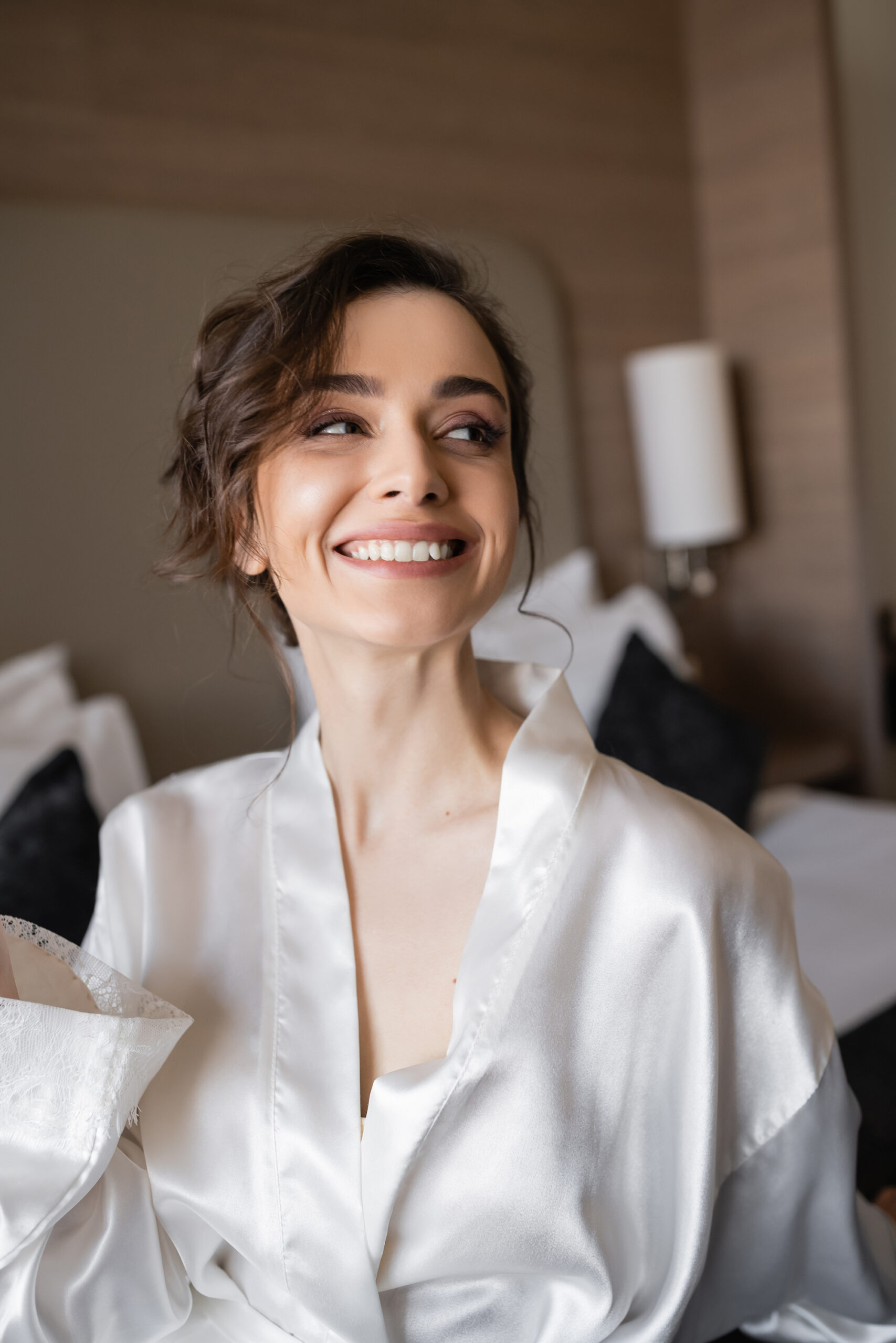 delightful woman with brunette hair and bridal makeup in white silk robe smiling and looking away in hotel suite on wedding day, special occasion