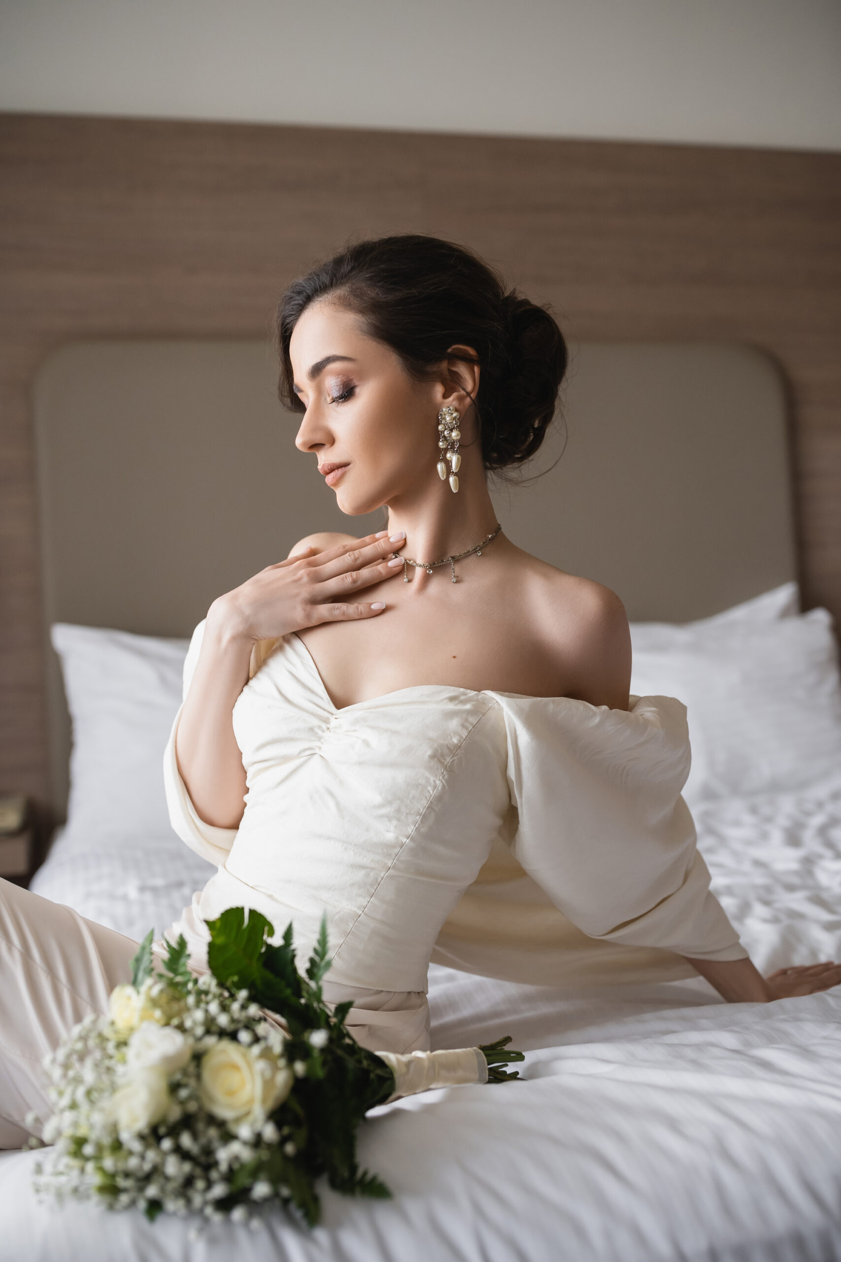 enchanting young bride in white dress and luxurious jewelry sitting on bed next to bridal bouquet with flowers in modern bedroom of hotel room on wedding day