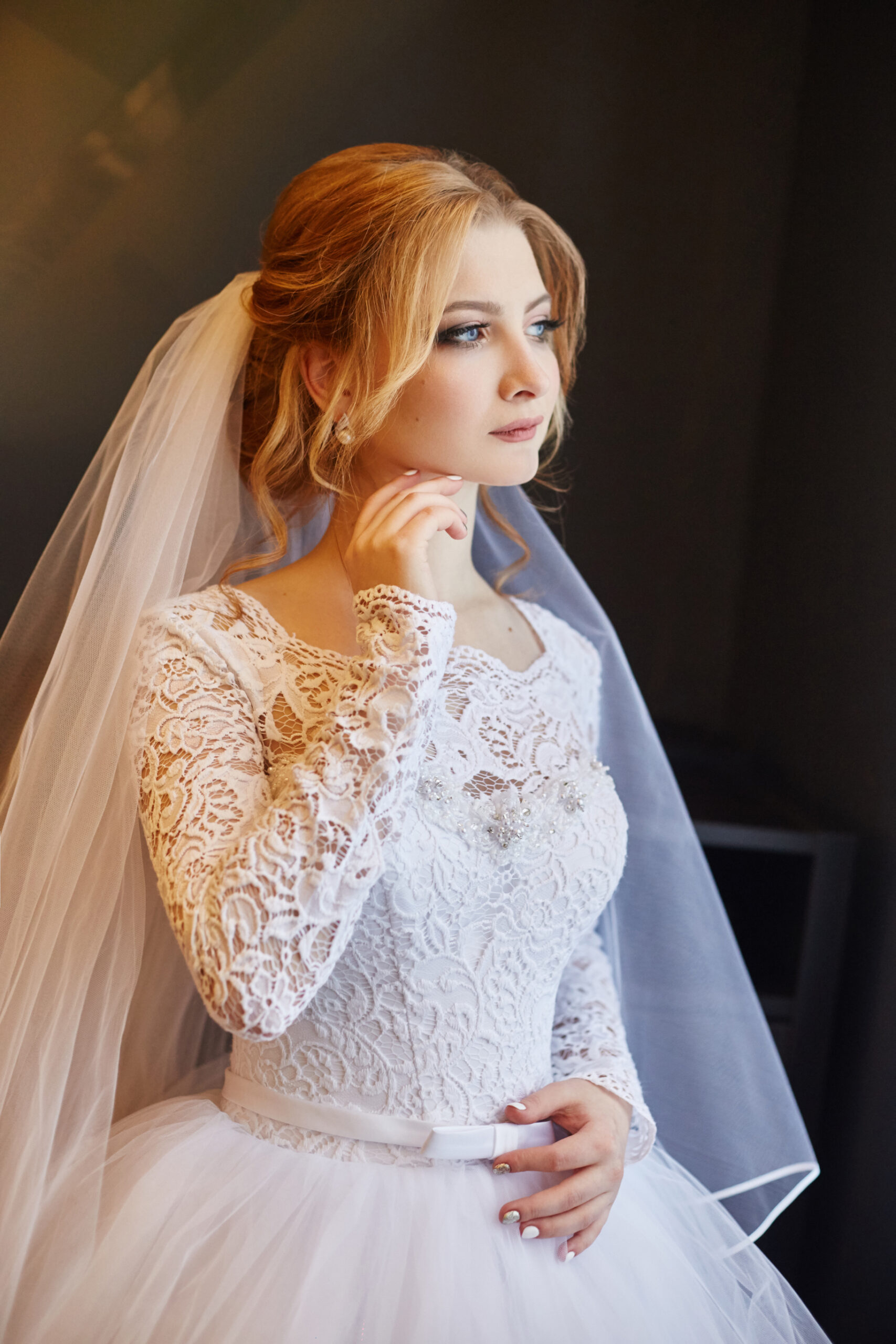 Portrait of a bride in a chic white wedding dress preparing for the wedding ceremony. Portrait of a blonde woman near a window in a white dress and a veil on her head. Bride is waiting for her groom