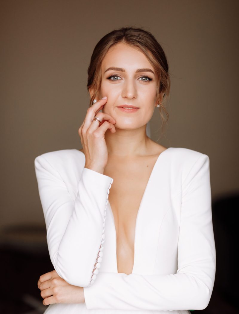 beautiful smiling young bride in long white satin dress with deep neckline, hairstyle and make up standing in hotel room. Trendy wedding style shoot. Wedding day. Bride's morning preparation