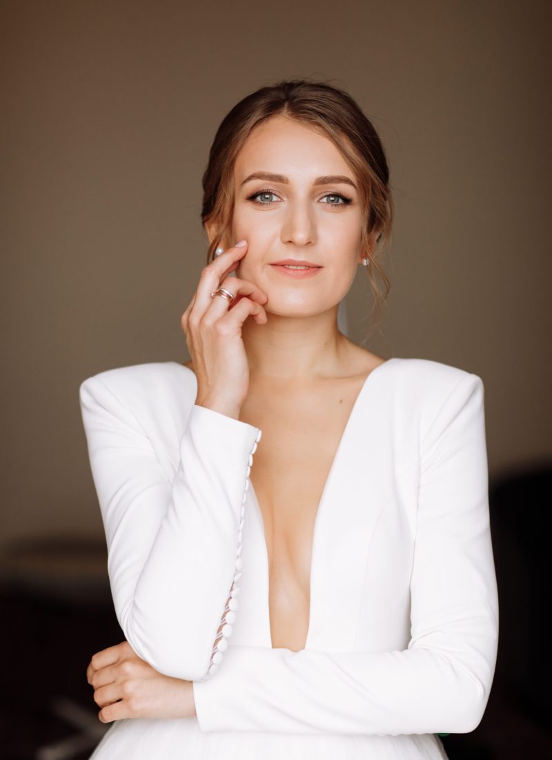 beautiful smiling young bride in long white satin dress with deep neckline, hairstyle and make up standing in hotel room. Trendy wedding style shoot. Wedding day. Bride's morning preparation