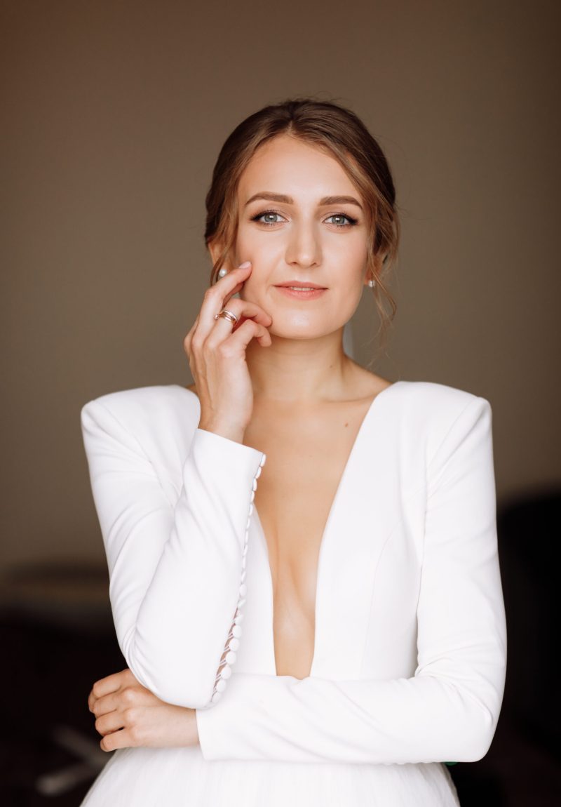 beautiful smiling young bride in long white satin dress with deep neckline, hairstyle and make up standing in hotel room. Trendy wedding style shoot. Wedding day. Bride's morning preparation
