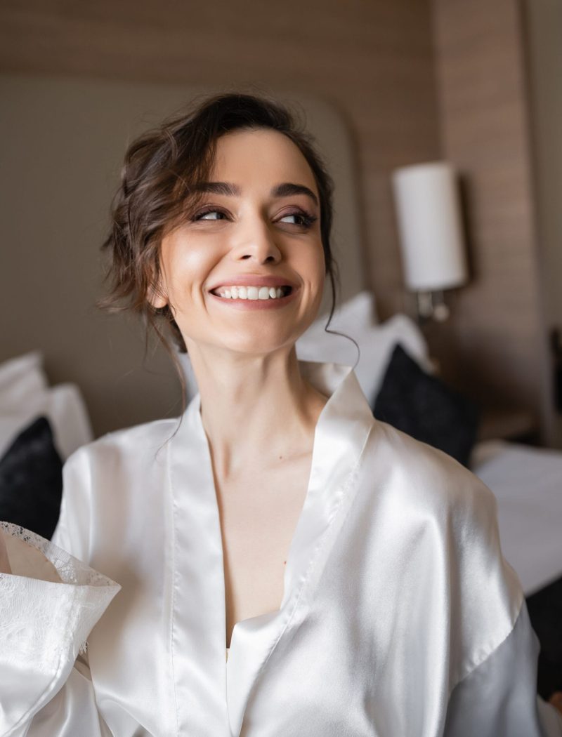 delightful woman with brunette hair and bridal makeup in white silk robe smiling and looking away in hotel suite on wedding day, special occasion