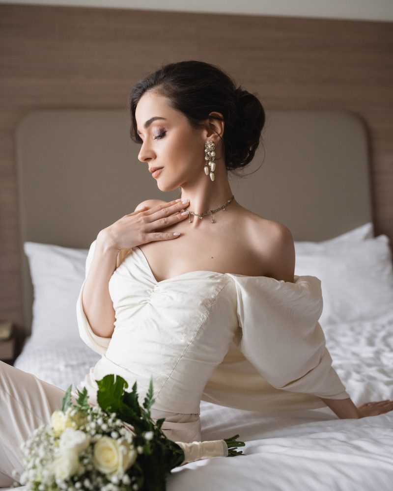 enchanting young bride in white dress and luxurious jewelry sitting on bed next to bridal bouquet with flowers in modern bedroom of hotel room on wedding day