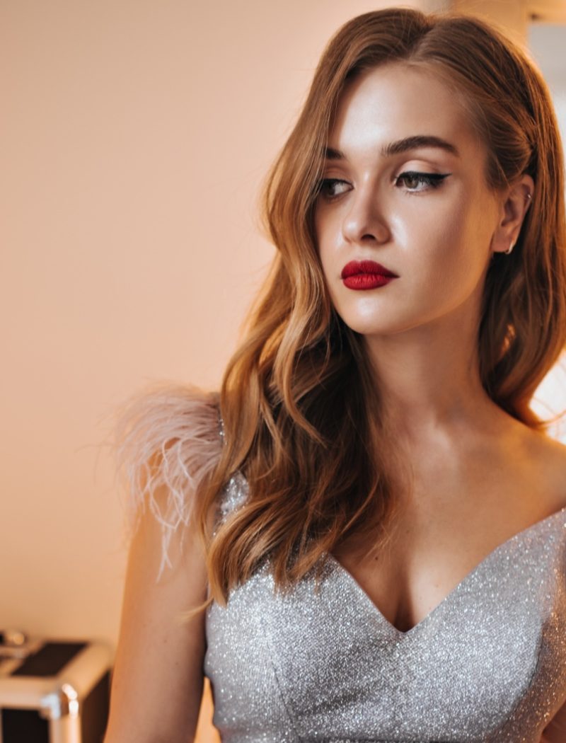Attractive lady in brilliant dress with feathers poses in dressing room. Portrait of blonde woman w