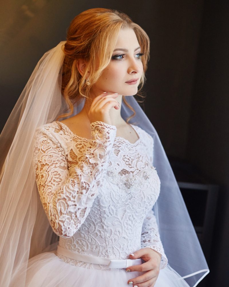 Portrait of a bride in a chic white wedding dress preparing for the wedding ceremony. Portrait of a blonde woman near a window in a white dress and a veil on her head. Bride is waiting for her groom