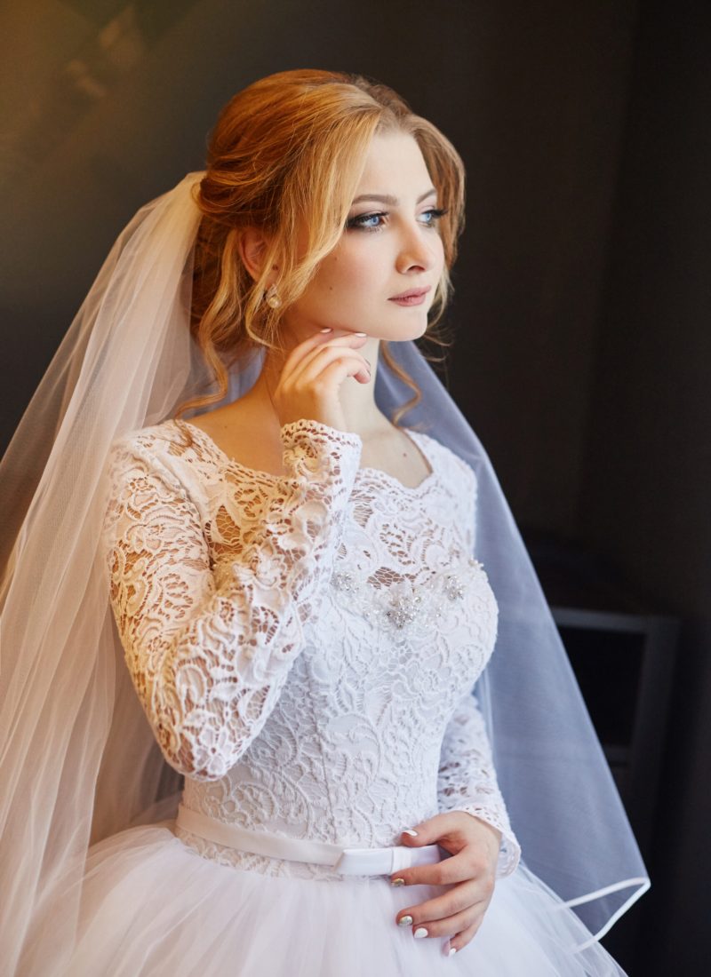 Portrait of a bride in a chic white wedding dress preparing for the wedding ceremony. Portrait of a blonde woman near a window in a white dress and a veil on her head. Bride is waiting for her groom