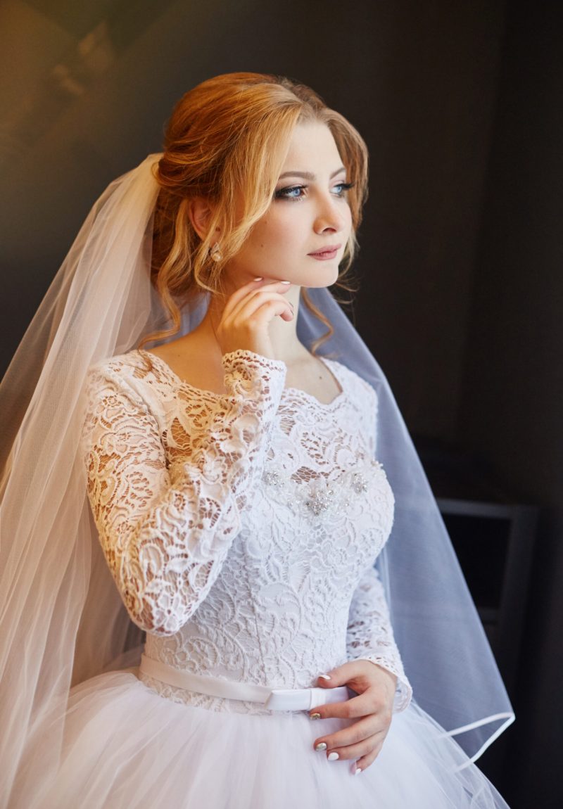Portrait of a bride in a chic white wedding dress preparing for the wedding ceremony. Portrait of a blonde woman near a window in a white dress and a veil on her head. Bride is waiting for her groom