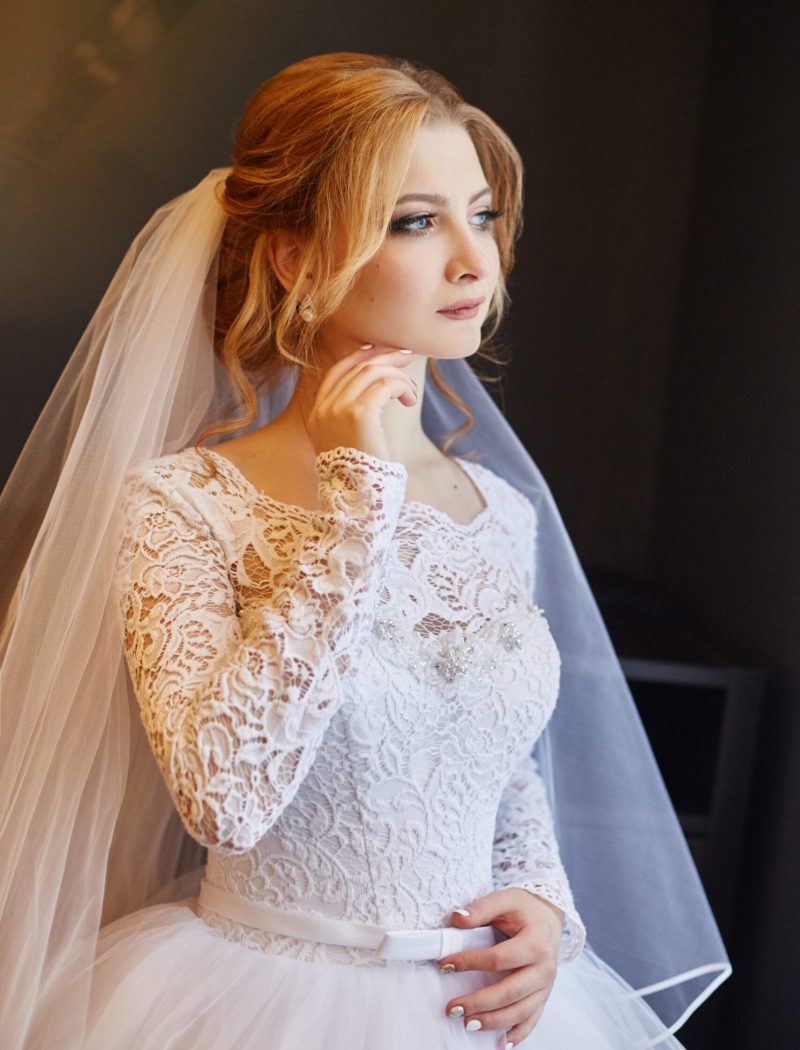 Portrait of a bride in a chic white wedding dress preparing for the wedding ceremony. Portrait of a blonde woman near a window in a white dress and a veil on her head. Bride is waiting for her groom